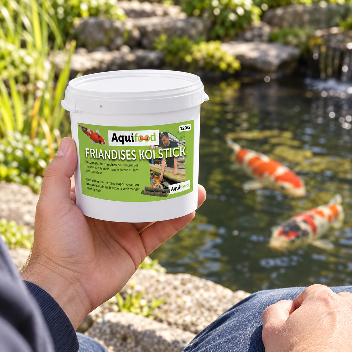 Mise en situation générée : présentation en main du pot Aquifood Koï Stick 120 g près du bassin.